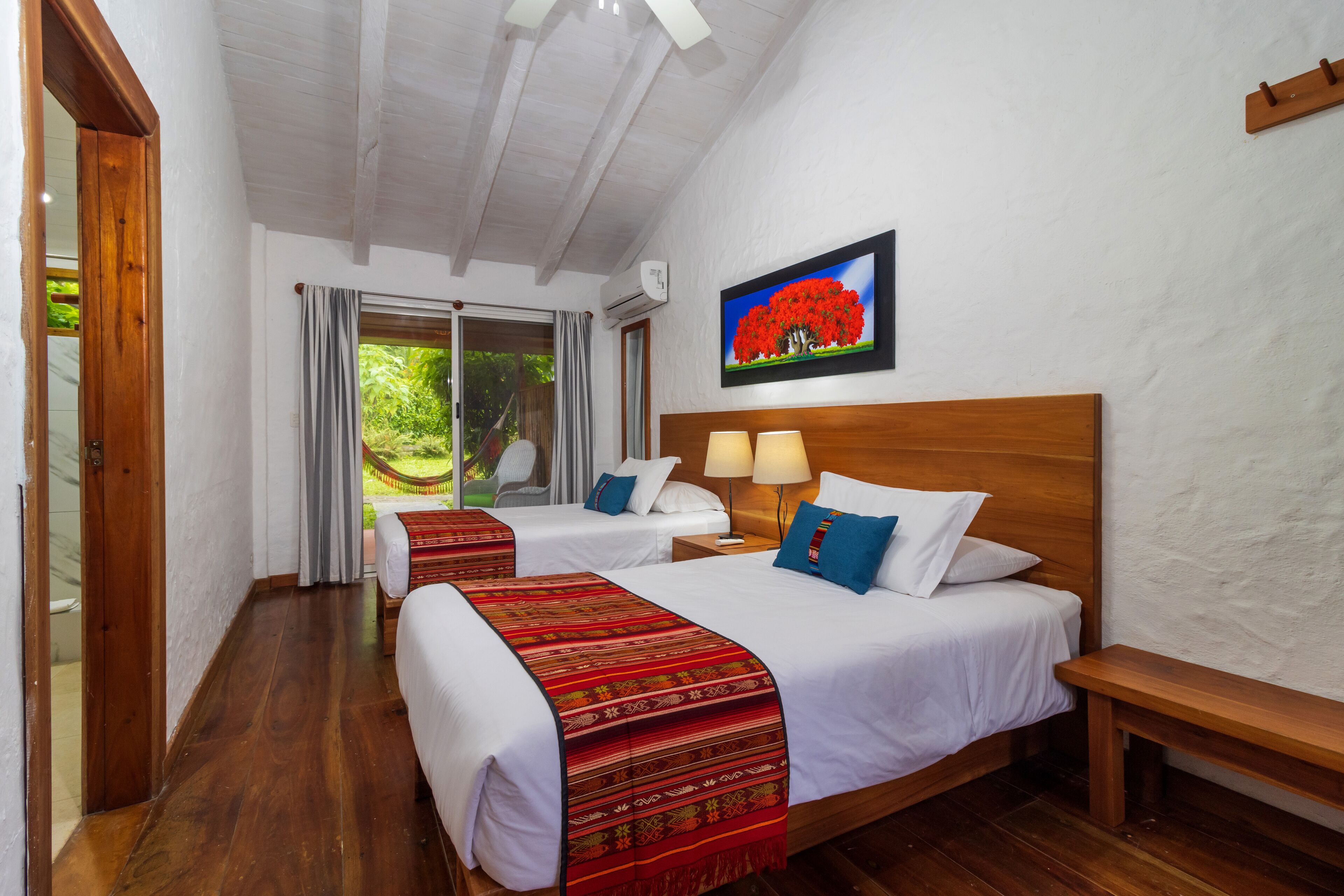 Habitación de hotel con dos camas, suelo de madera, una banca, televisor en la pared y ventana con cortinas.