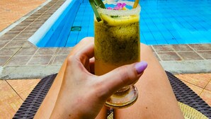 Una mano sosteniendo una bebida con adorno verde frente a una piscina con borde de baldosas y una zona de estar cubierta.