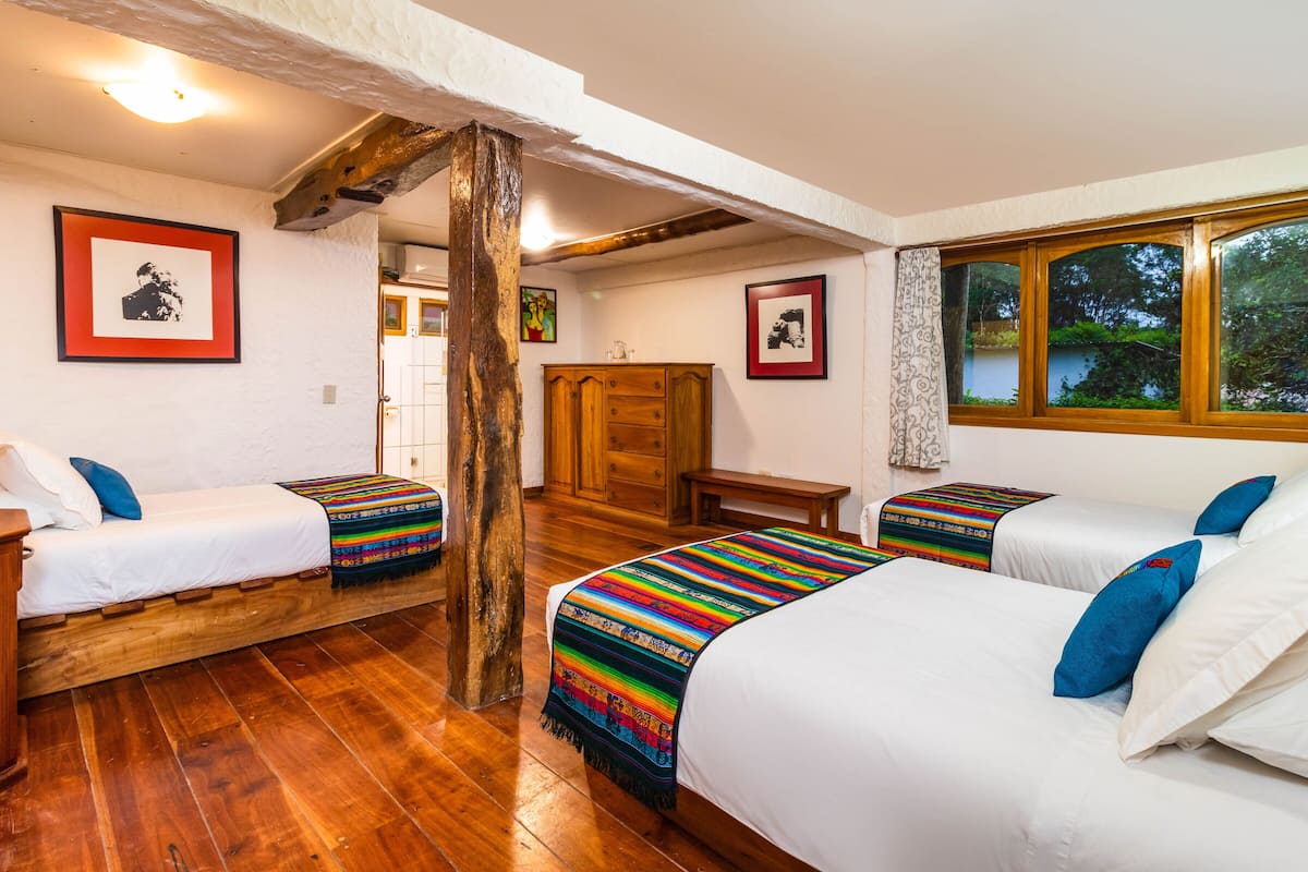 A room with two beds, wooden flooring, a wooden wardrobe, and a window with a view of greenery.