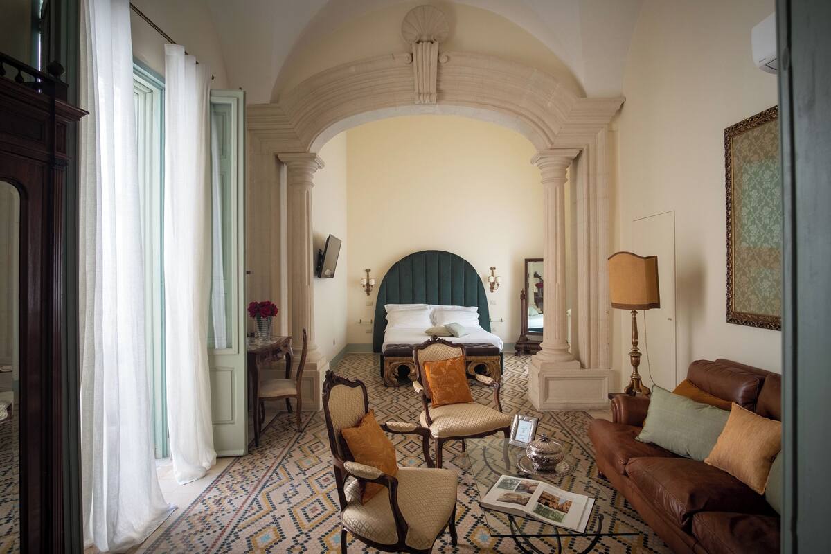 Habitación de hotel con una cama grande, zona de estar con sofá y sillones, suelo estampado y un arco de entrada.