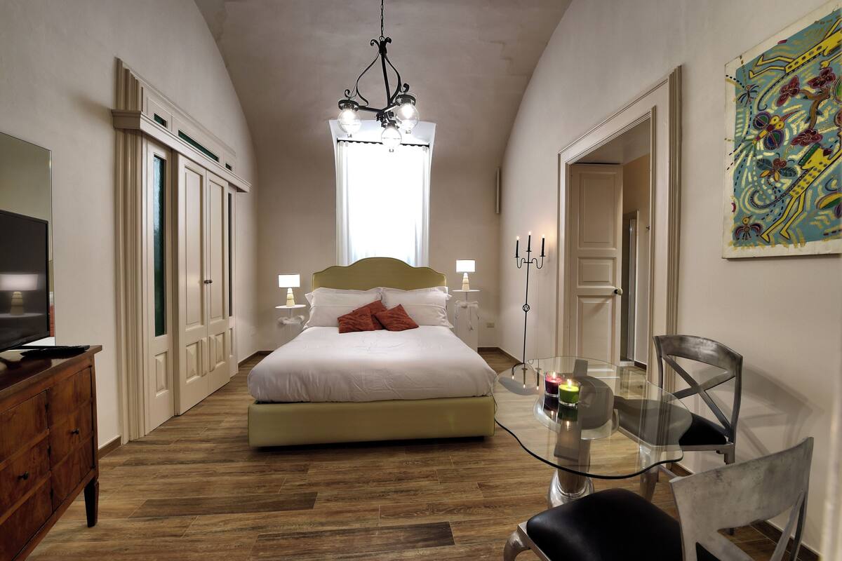 Un dormitorio con cama, mesitas de noche, televisor, una mesa de vidrio con sillas y una araña.