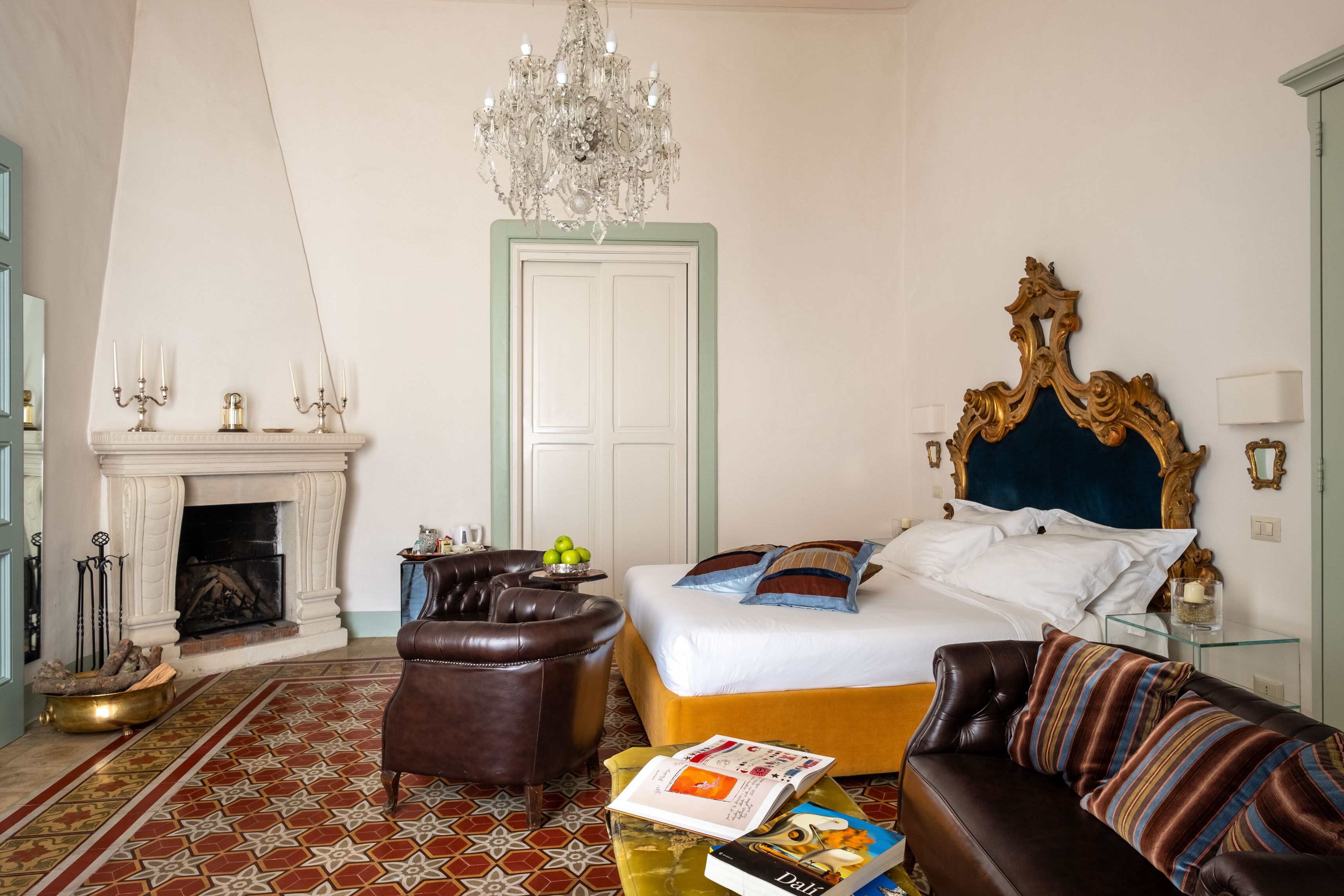 A bedroom with a bed, a leather armchair, a sofa, a fireplace, and a chandelier.