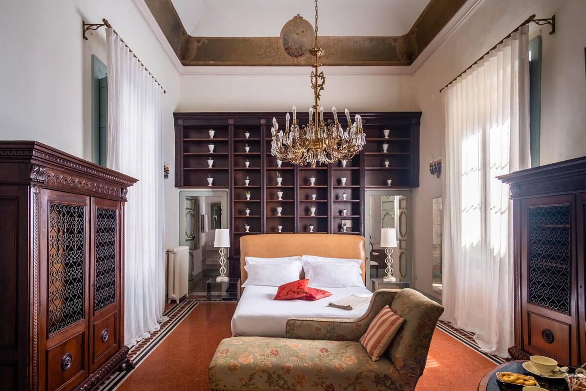 Un dormitorio con una cama grande, un candelabro, un sofá floral y una estantería llena de objetos decorativos.