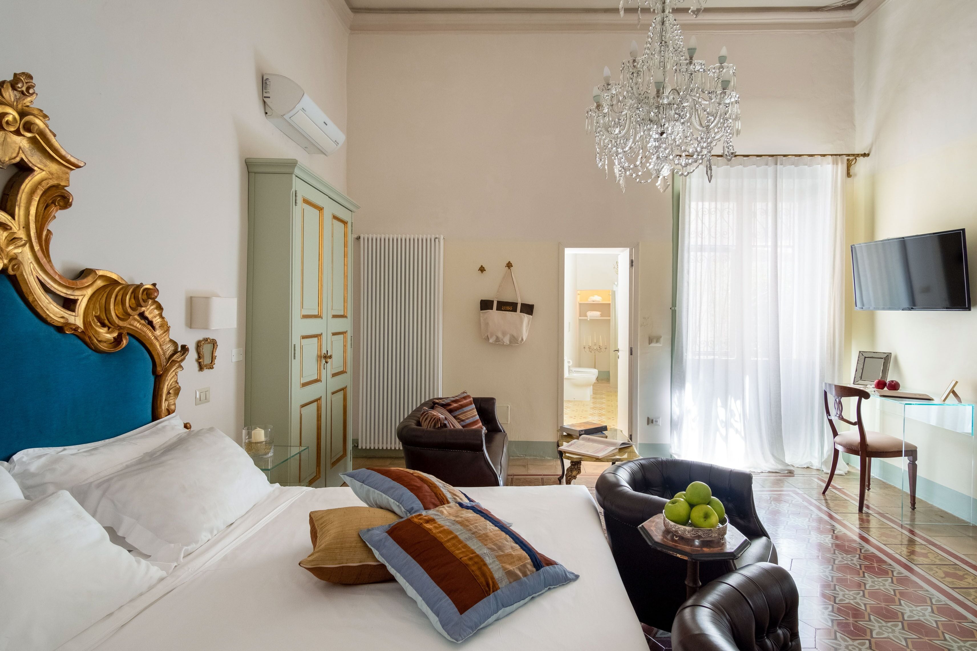 A hotel room with a large bed, a chandelier, a television, and a view of the bathroom.