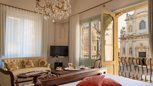 A hotel room with a bed, a sofa, a TV, a dining table with a tray of food, and a view of a historic building. - Palazzo Guido Boutique Hotel (Lecce)