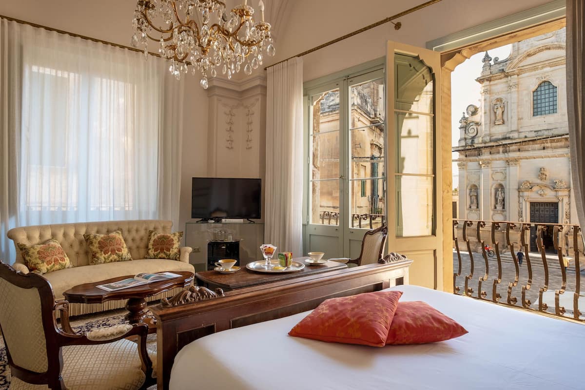 Habitación de hotel con cama, sofá, televisor, mesa de comedor y una bandeja de comida, y vista a un edificio histórico.