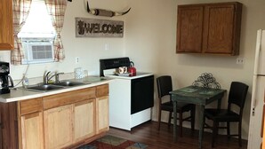 A compact kitchen with wooden cabinets, a white stove, and a dining area with a table and chairs. - Tumble Weed Cabin of Lonesome Wind Ranch (Floresville)