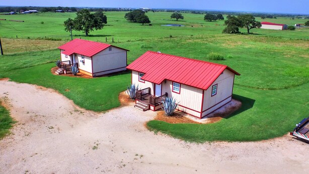 Two small, single-story buildings with red roofs, surrounded by green fields and trees, with a dirt path leading to them. - Tumble Weed Cabin of Lonesome Wind Ranch (Floresville)