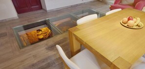 Un espace repas avec une table en bois et des chaises blanches, une cloison en verre et un mur en pierre éclairé d’une lumière chaleureuse.