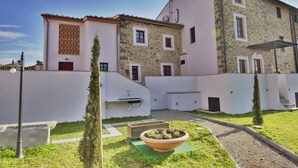 Un bâtiment à deux étages, doté d’une façade en pierre et d’un toit plat. On y trouve un petit jardin avec un grand bac à fleurs et un chemin menant à l’entrée.