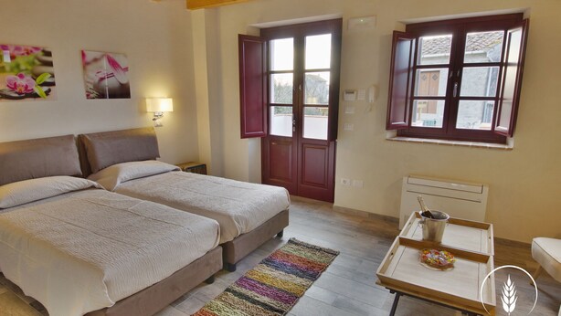 Une chambre avec deux lits, un plafond en bois, une fenêtre avec des volets rouges, un radiateur et une petite table sur laquelle se trouvent une bouteille et des verres.