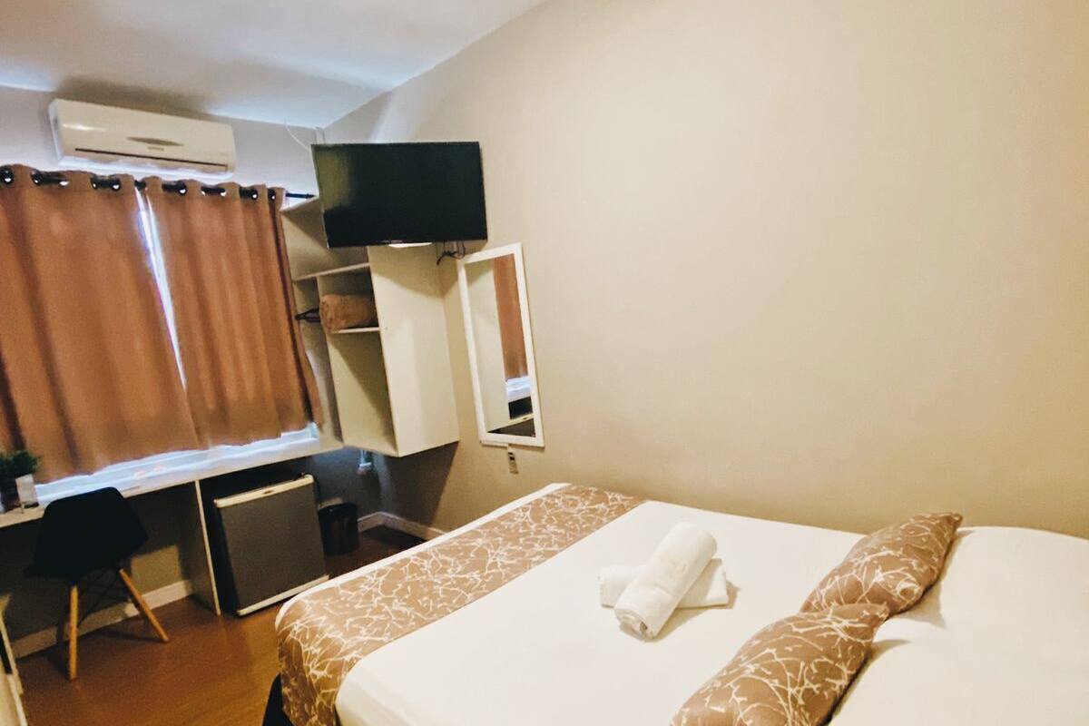 A hotel room with a bed, a desk, a chair, a television, and an air conditioning unit.