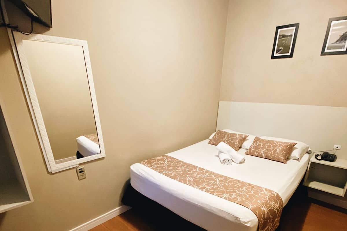 A hotel room with a large bed, a bedside table, a mirror, and a wall-mounted TV.