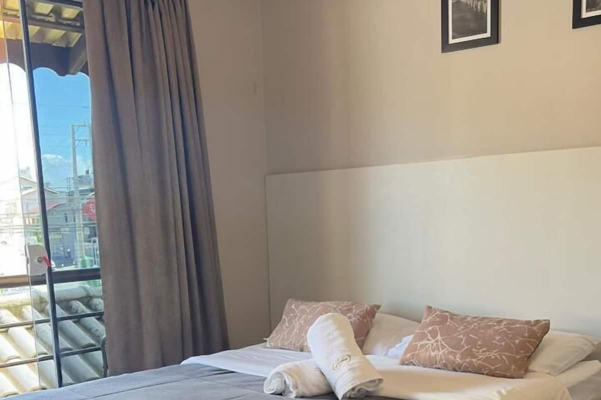 A hotel room with a bed, a window with curtains, an air conditioning unit, and two framed pictures on the wall.
