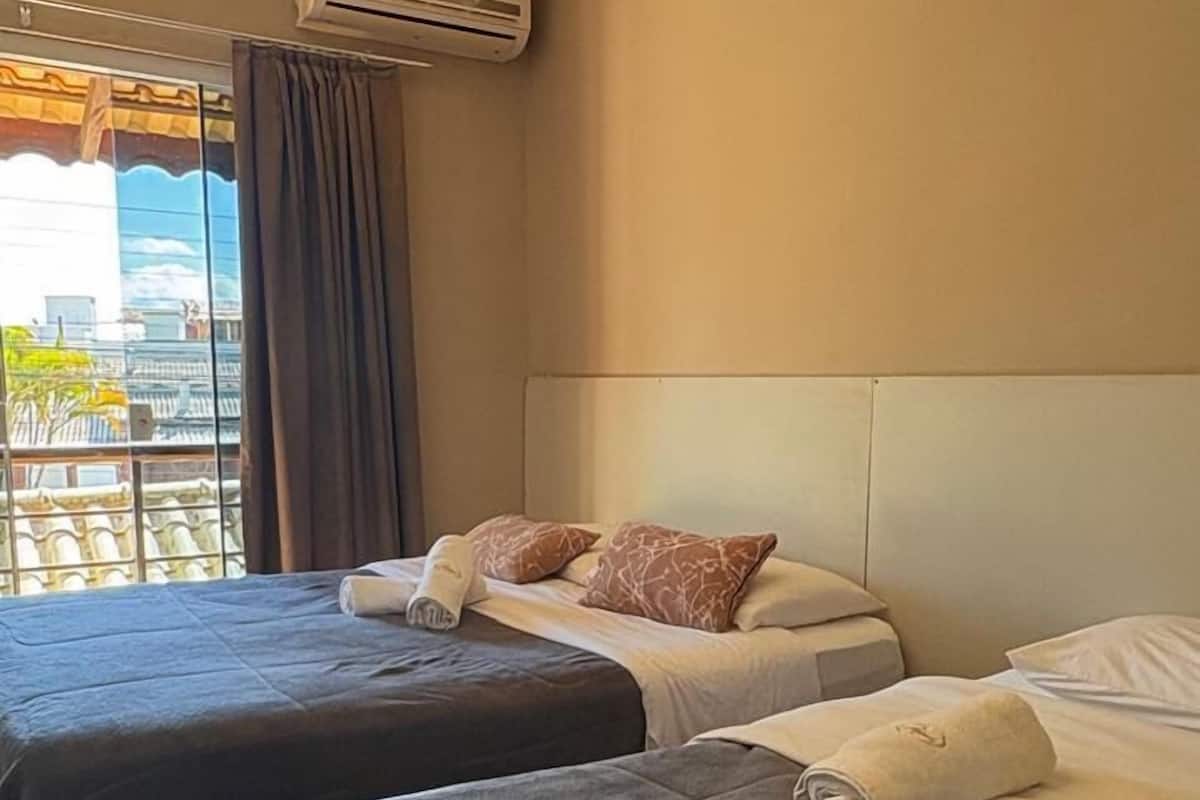A hotel room with two beds, a large window with curtains, an air conditioning unit, and a view of buildings outside.