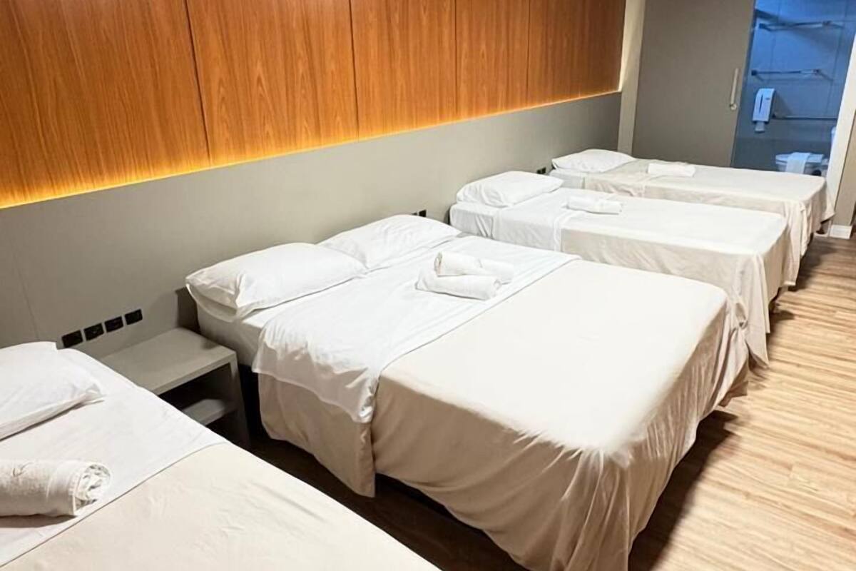 A hotel room with four beds arranged in a row, a wooden headboard, a ceiling-mounted air conditioning unit, and a view of the outside through a window.