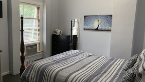 Quarto com cama, cômoda, janela com persianas e uma obra de arte na parede.