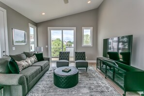 A modern living room with a grey sofa, two armchairs, a round ottoman, a television, and a large window with a view of the outdoors.
