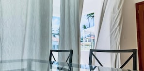 A glass-top table with a geometric metal frame, a white curtain, and a view of buildings and palm trees outside.