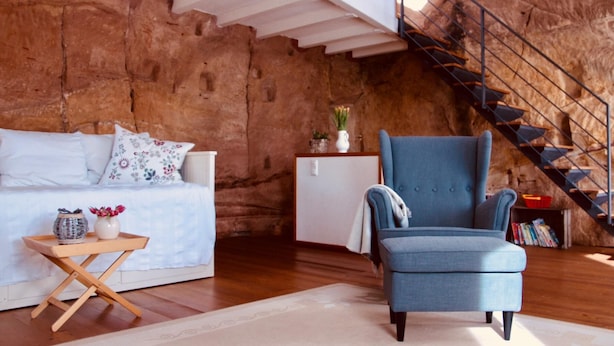 A room with a rustic stone wall, a blue armchair, a white sofa, a small table with decorative items, and a staircase with a metal railing. - FELSENHAUS with view over Trier - living in the rock (Trier)