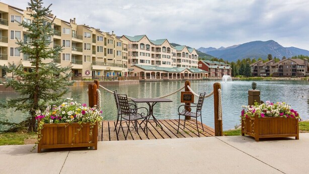 A lakeside deck with a table and chairs, overlooking a body of water and a row of buildings with a mountain backdrop. - 1546 | Wonderful View, Pool & Spa On Keystone Lake (Keystone)
