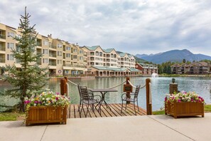 A lakeside deck with a table and chairs, overlooking a body of water and a row of buildings with a mountain backdrop.