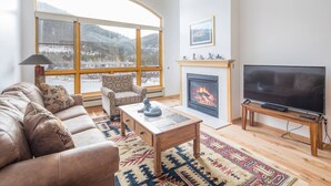 A living room with a sofa, armchair, fireplace, TV, and a large window with a view of mountains. - 1546 | Wonderful View, Pool & Spa On Keystone Lake (Keystone)