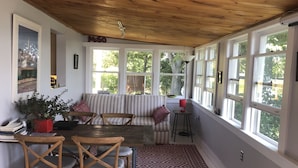 Una habitación acogedora con techo de madera, un sofá a rayas, una mesa de comedor con sillas y ventanas grandes que ofrecen vistas al verde exterior.