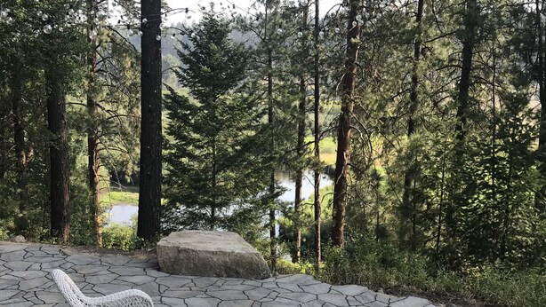 Une terrasse en pierre avec des chaises en osier et un foyer, entourée d’une forêt et d’un lac.