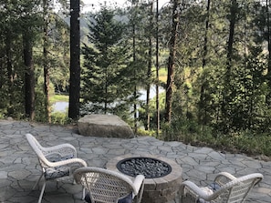 A stone patio with wicker chairs and a fire pit, surrounded by a forest and a lake.