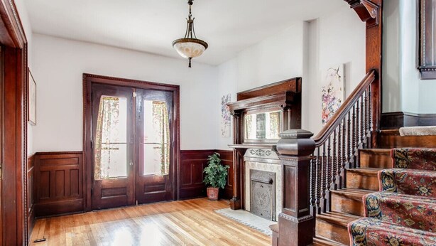 A room with wooden flooring, a fireplace, a staircase with ornate railings, and a door with floral curtains. - Beautiful Victorian Louisville Home (Louisville)