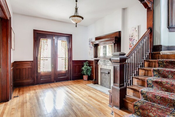 A room with wooden flooring, a fireplace, a staircase with ornate railings, and a door with floral curtains.
