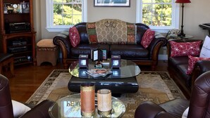 Un salon avec un plafond haut, une grande fenêtre donnant sur les arbres, un canapé en cuir noir, une table basse en verre et une bibliothèque.