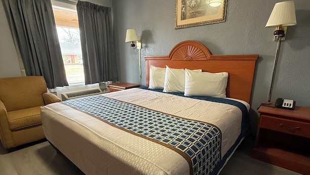A hotel room with a large bed, a wooden headboard, two bedside lamps, a chair, and a painting on the wall.
