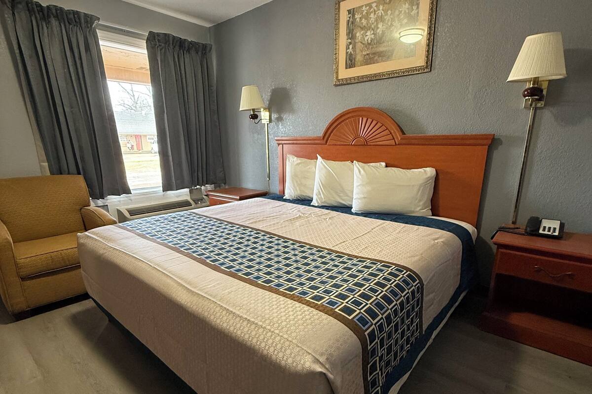 A hotel room with a large bed, a wooden headboard, two bedside lamps, a chair, and a painting on the wall.