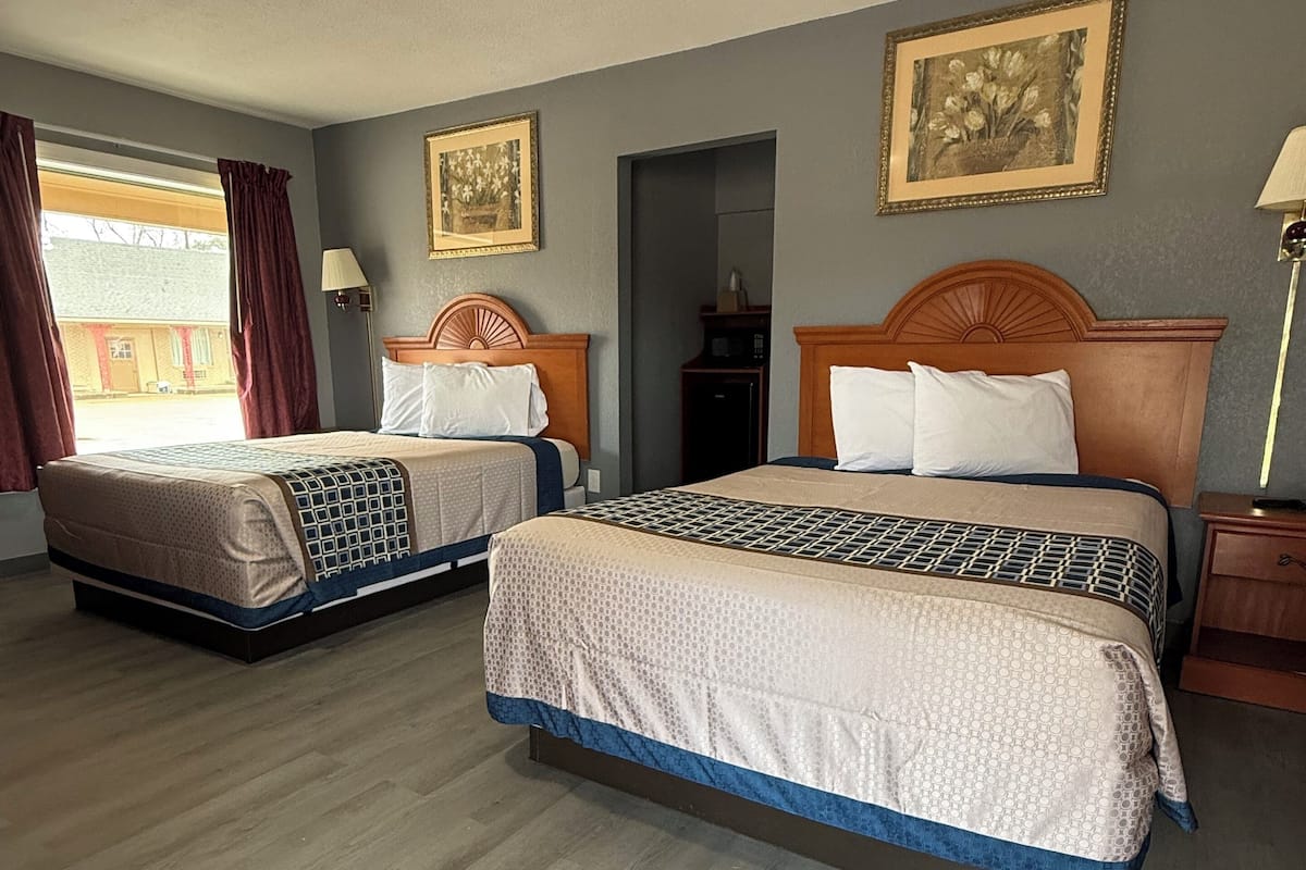A hotel room with two beds, a wooden headboard, a ceiling light, a framed picture on the wall, and a window with curtains.