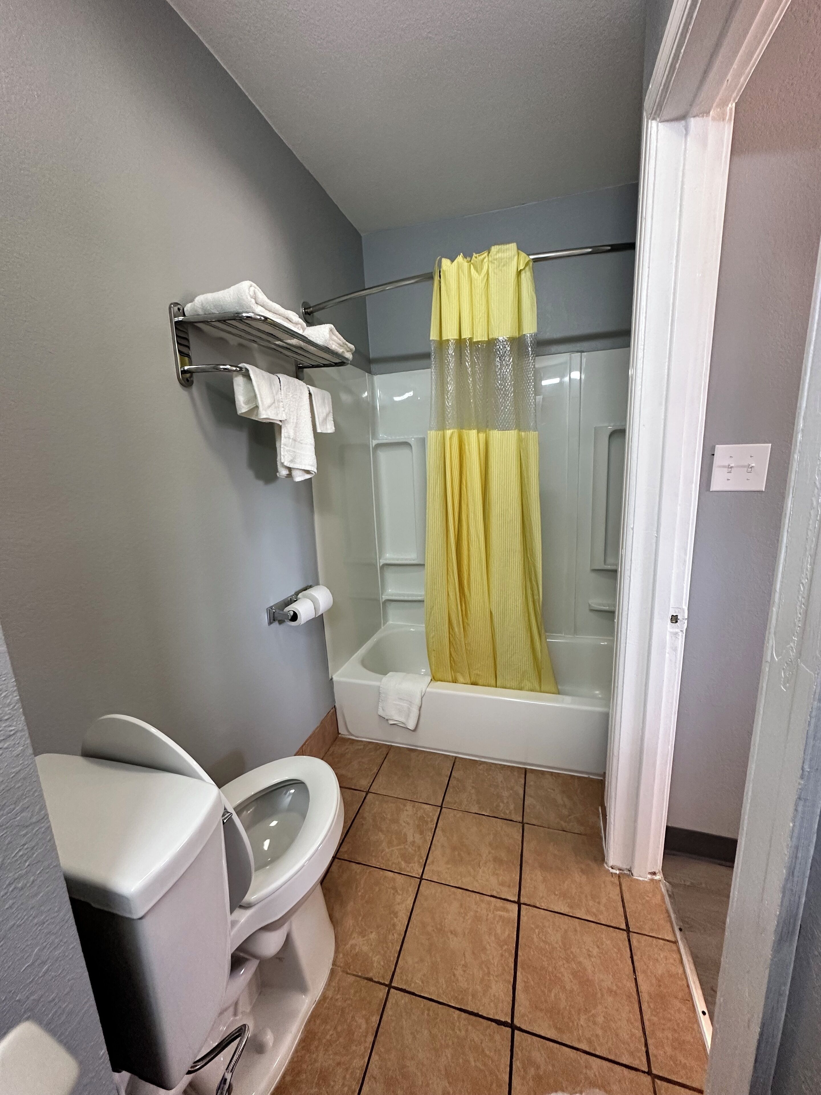 A bathroom with a toilet, a shower with a yellow curtain, a towel rack with white towels, and a white bathtub.