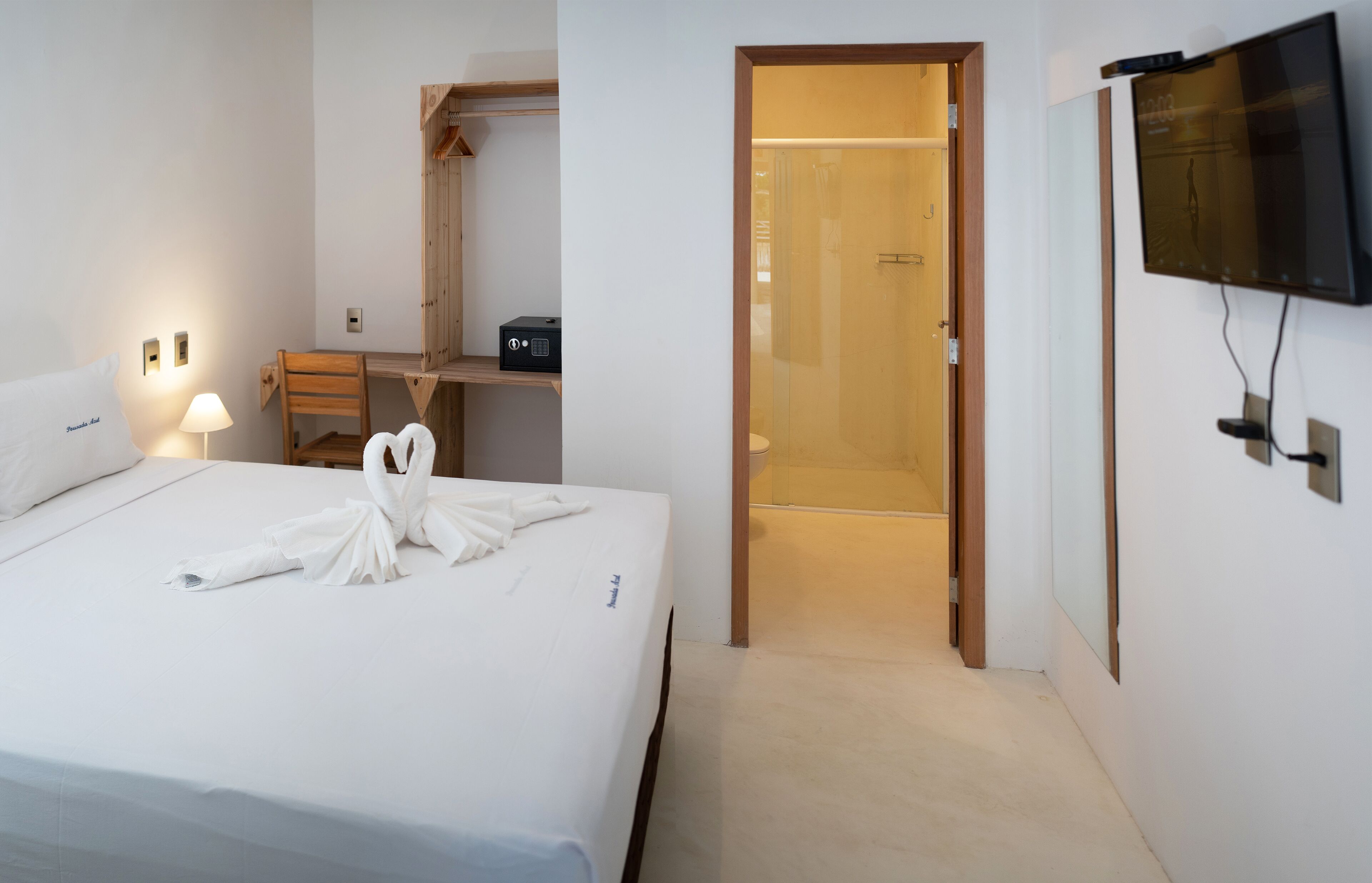 Habitación de hotel con una cama bien tendida, un escritorio con silla, un televisor en la pared y un espacio con ducha.