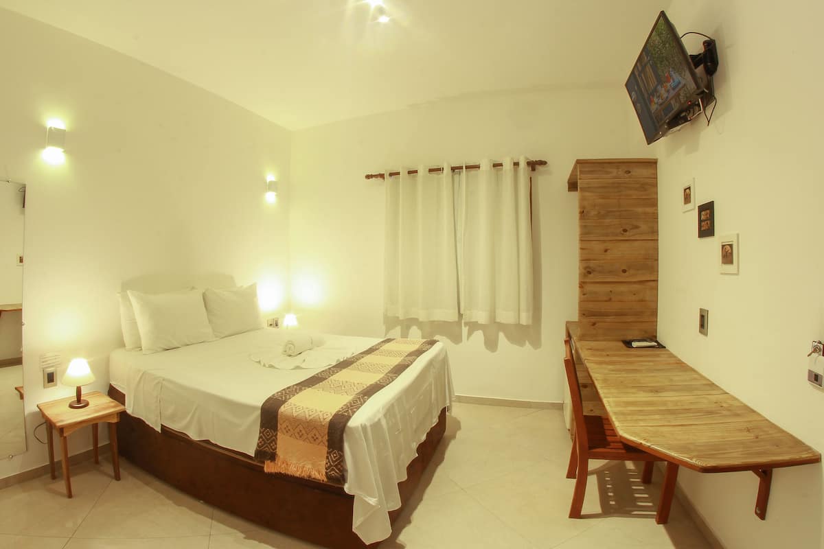 A hotel room with a bed, bedside table, lamp, wooden desk, and curtains.