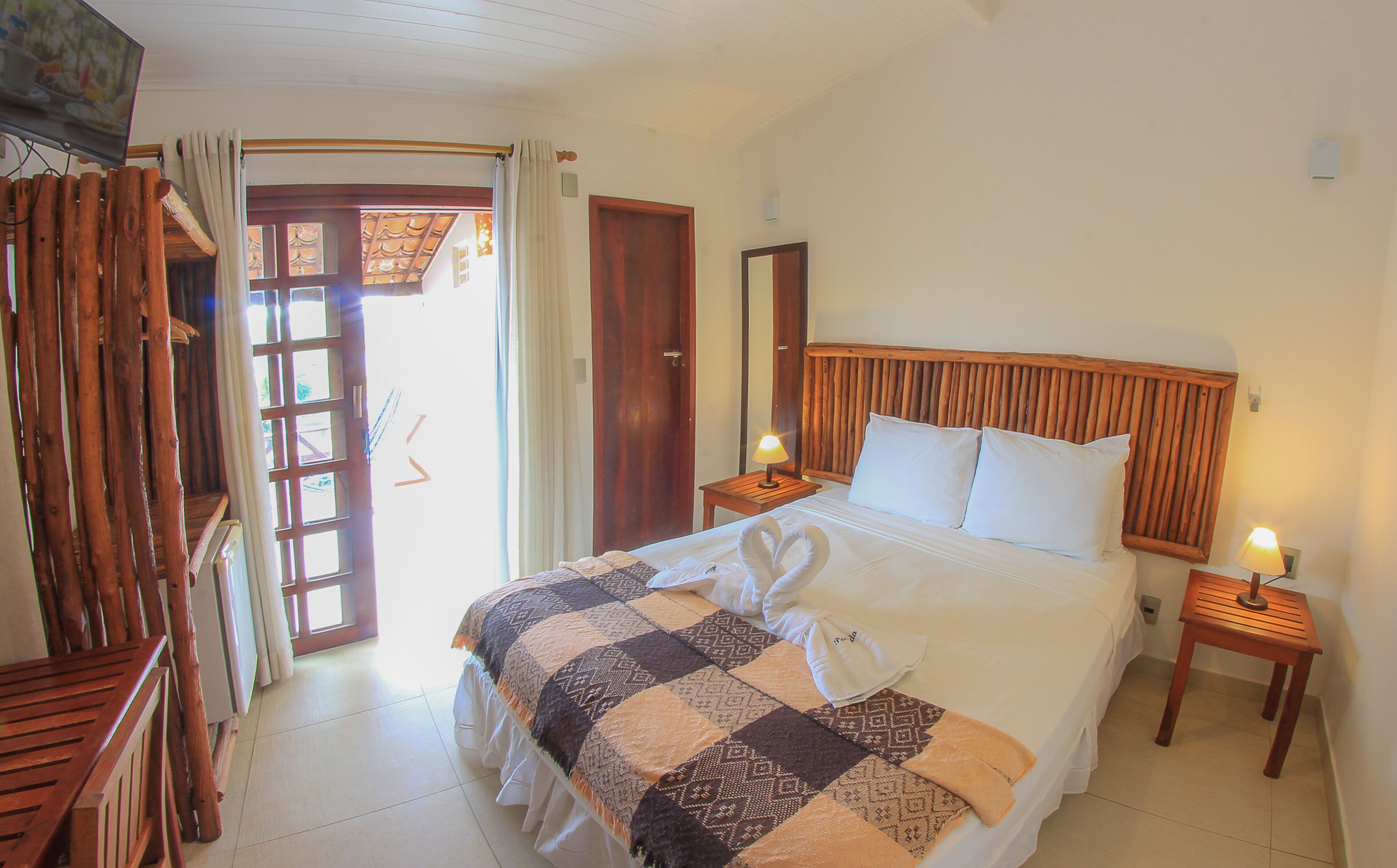 A hotel room with a bed, a wooden headboard, two bedside tables with lamps, a wooden bench, a large window with curtains, and a door leading outside.