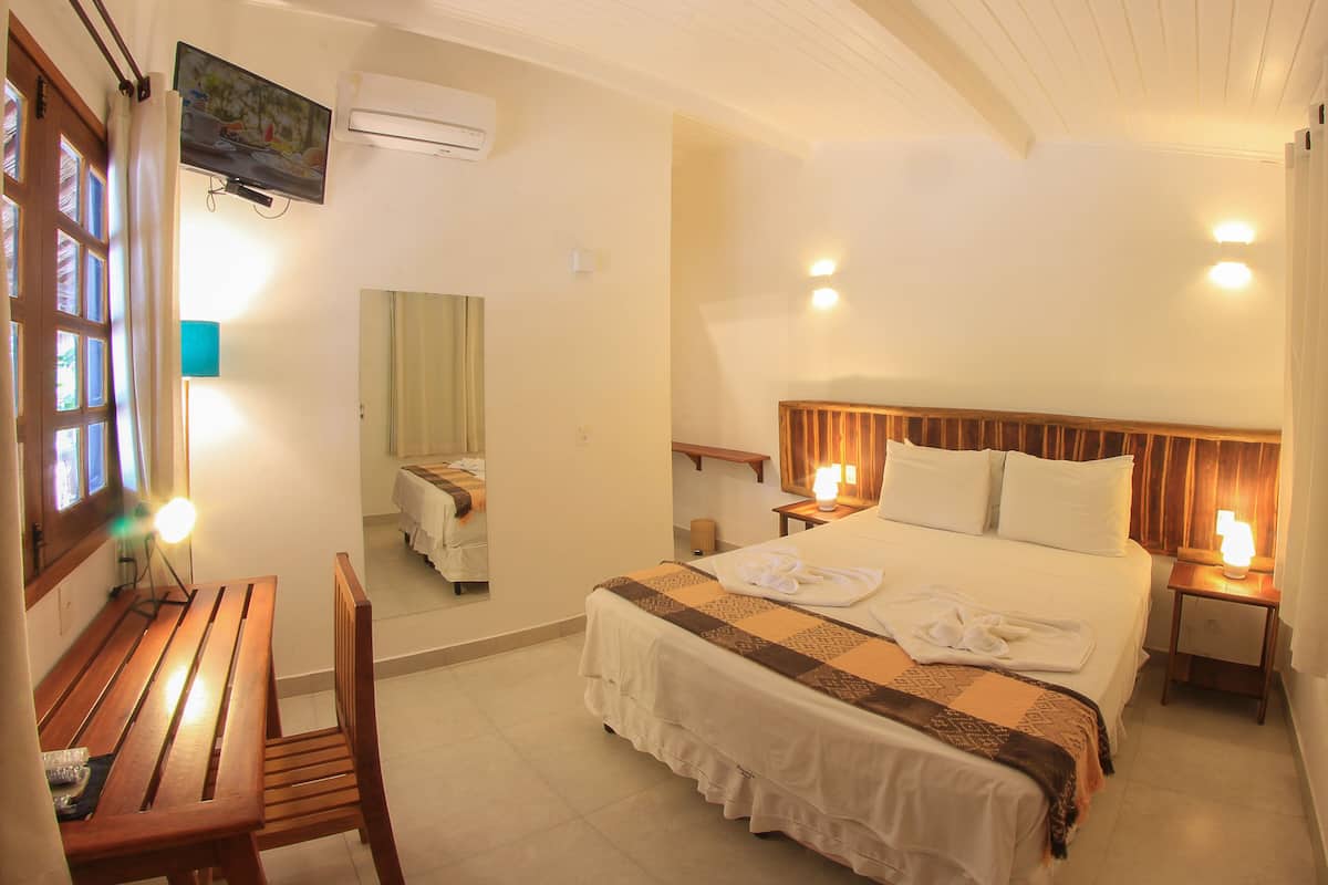A hotel room with a bed, bedside tables, a wooden bench, a mirror, and an air conditioning unit.