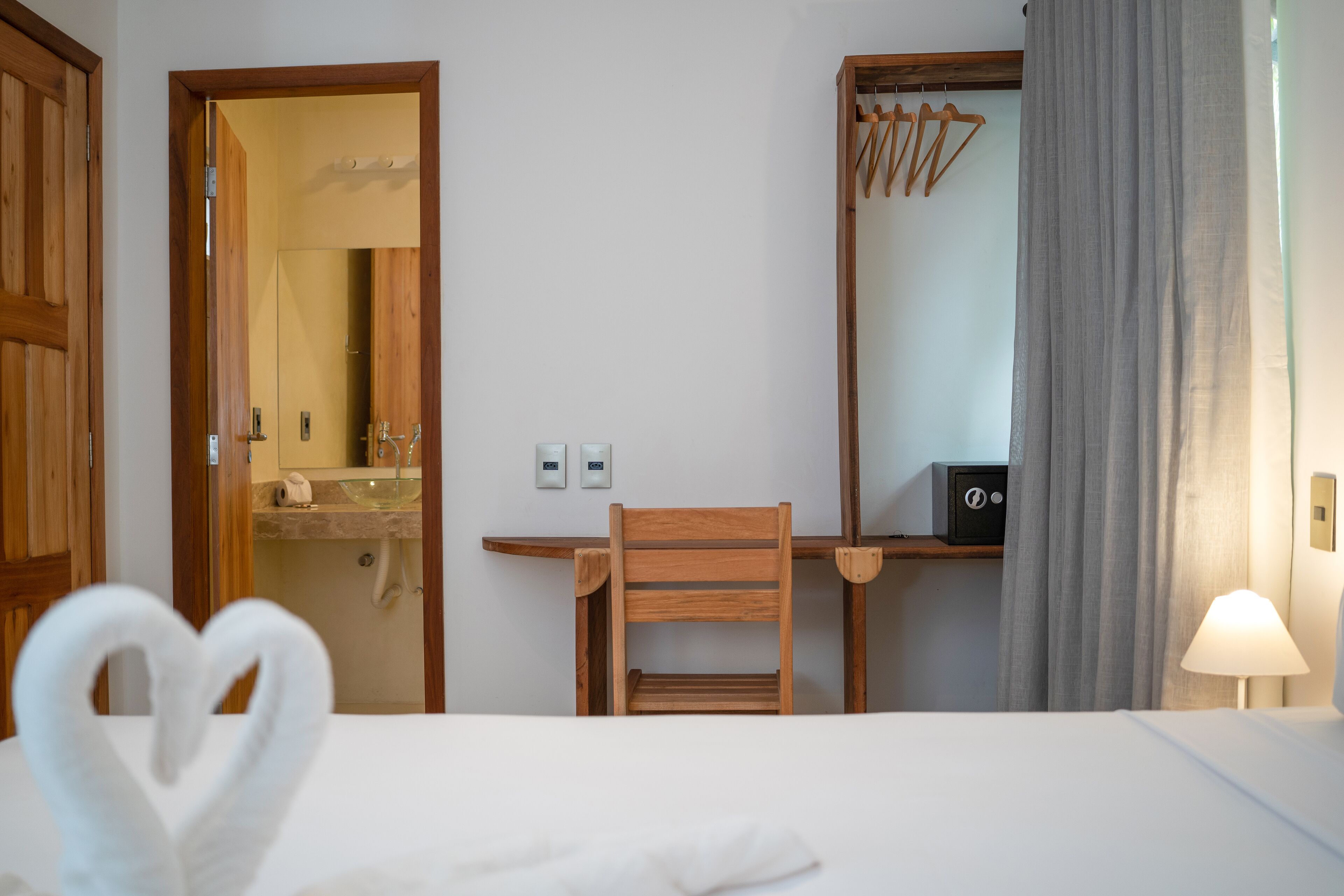 A hotel room with a bed, a wooden chair, a desk with a mirror, a lamp, and a bathroom visible through an open door.