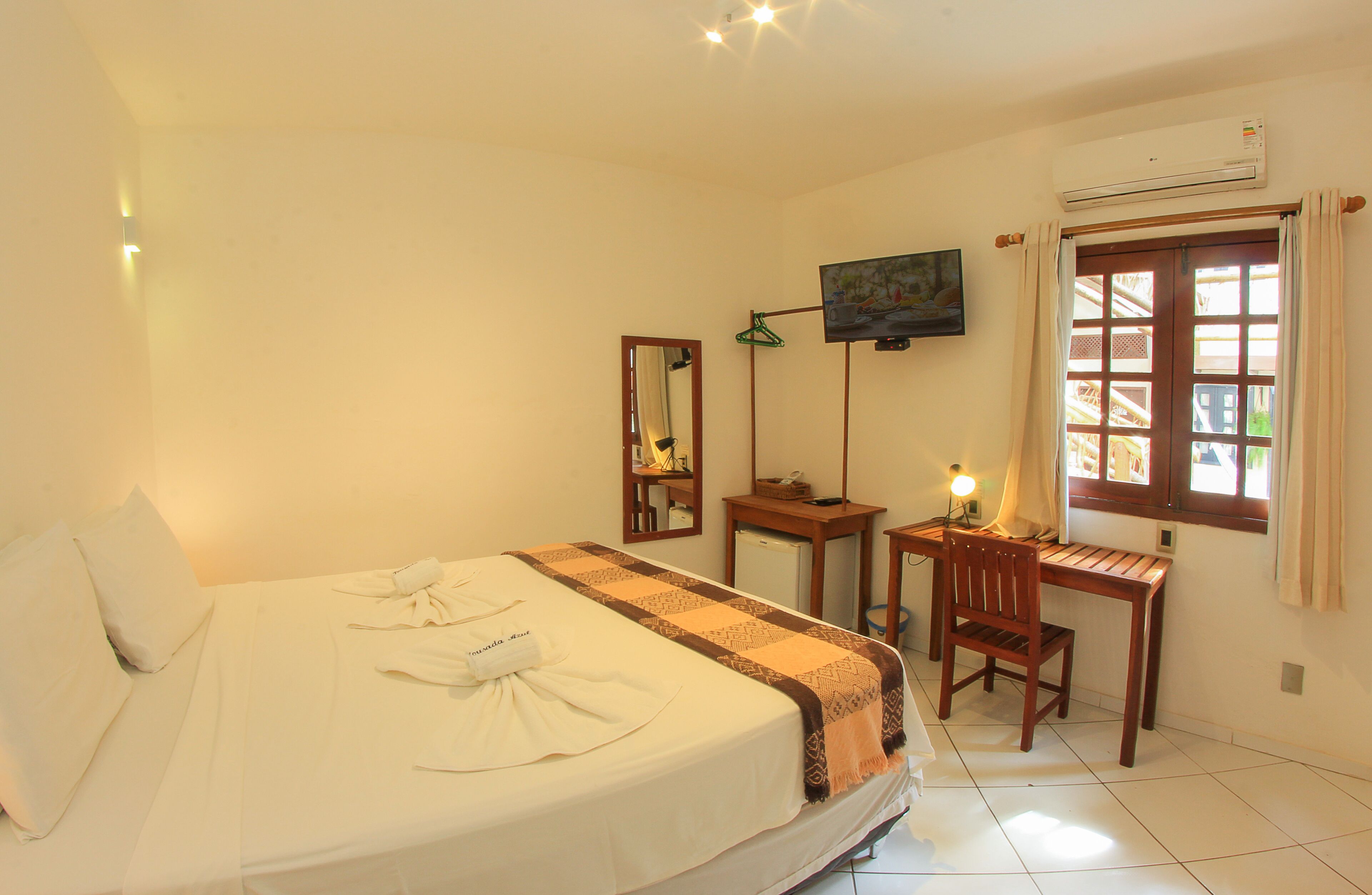 A hotel room with a bed, a desk, a chair, a TV, and an air conditioning unit.