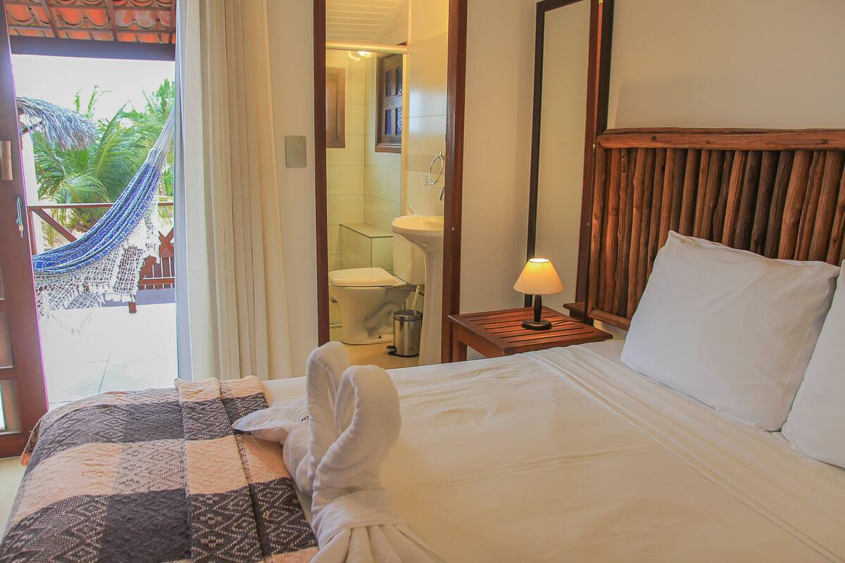 A hotel room with a bed, a nightstand with a lamp, a bathroom visible through an open door, and a hammock outside.