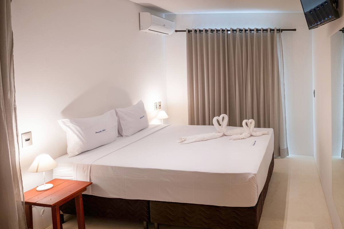 A hotel room with a bed, a bedside table with a lamp, a curtain, and a wall-mounted air conditioner.
