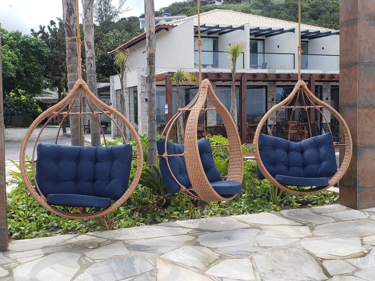 Three wicker swing chairs with blue cushions in a garden area with a stone floor, surrounded by greenery and a modern building in the background.