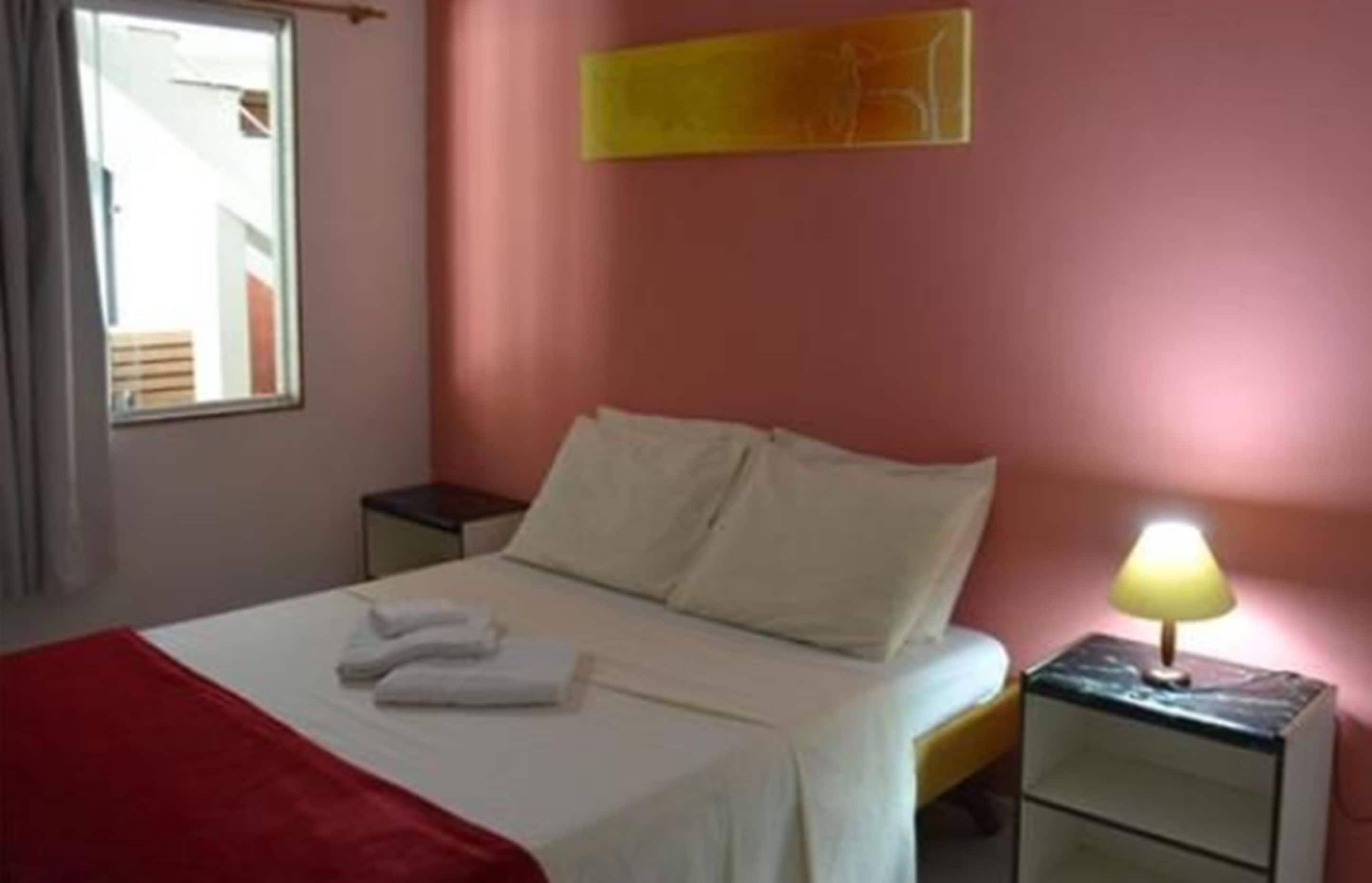 A hotel room with a bed, a nightstand with a lamp, a window, and a red blanket on the bed.