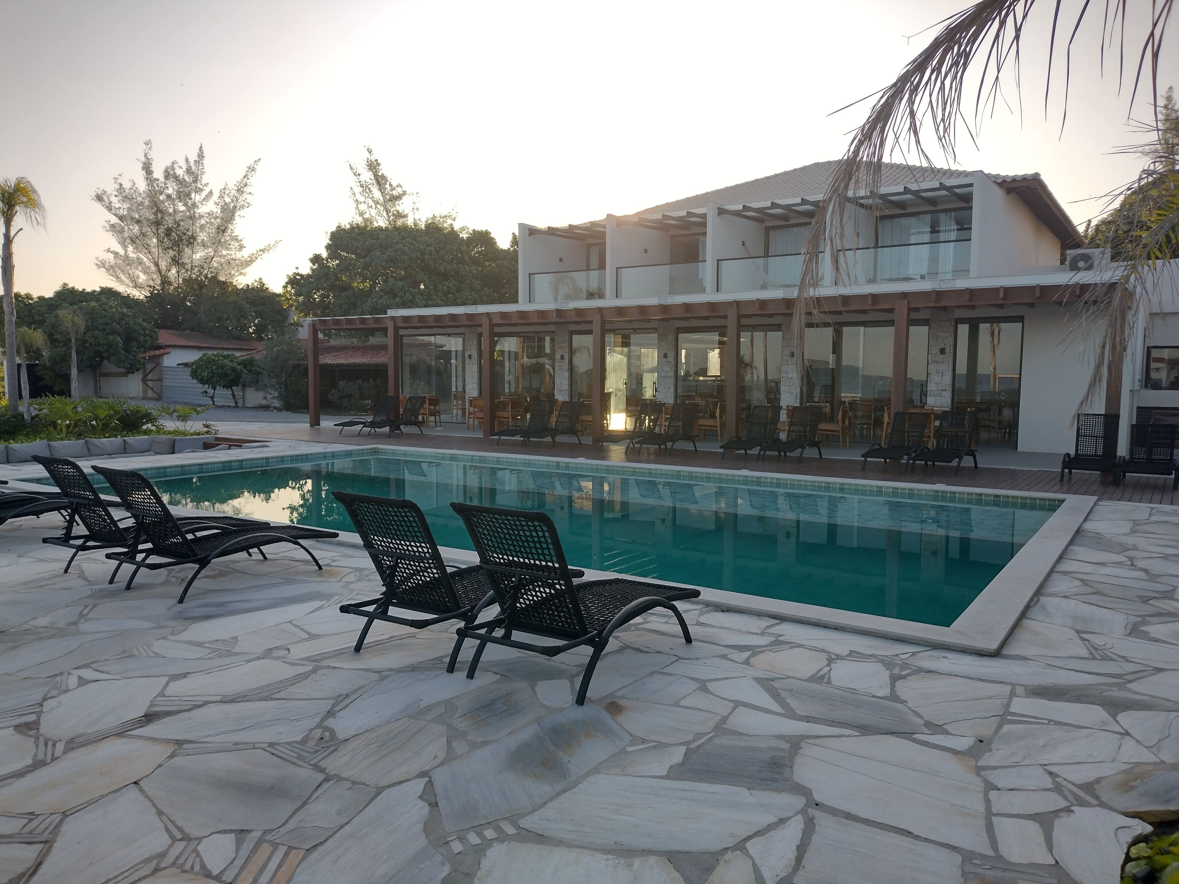 Una piscina con sillones de descanso, un área de estar techada al aire libre y un edificio moderno con grandes ventanales.