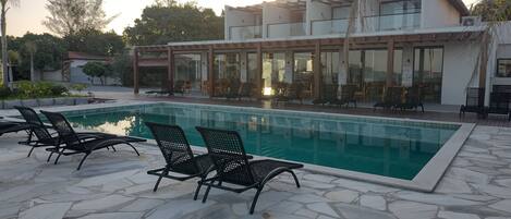 Una piscina con sillones de descanso, un área de estar techada al aire libre y un edificio moderno con grandes ventanales.