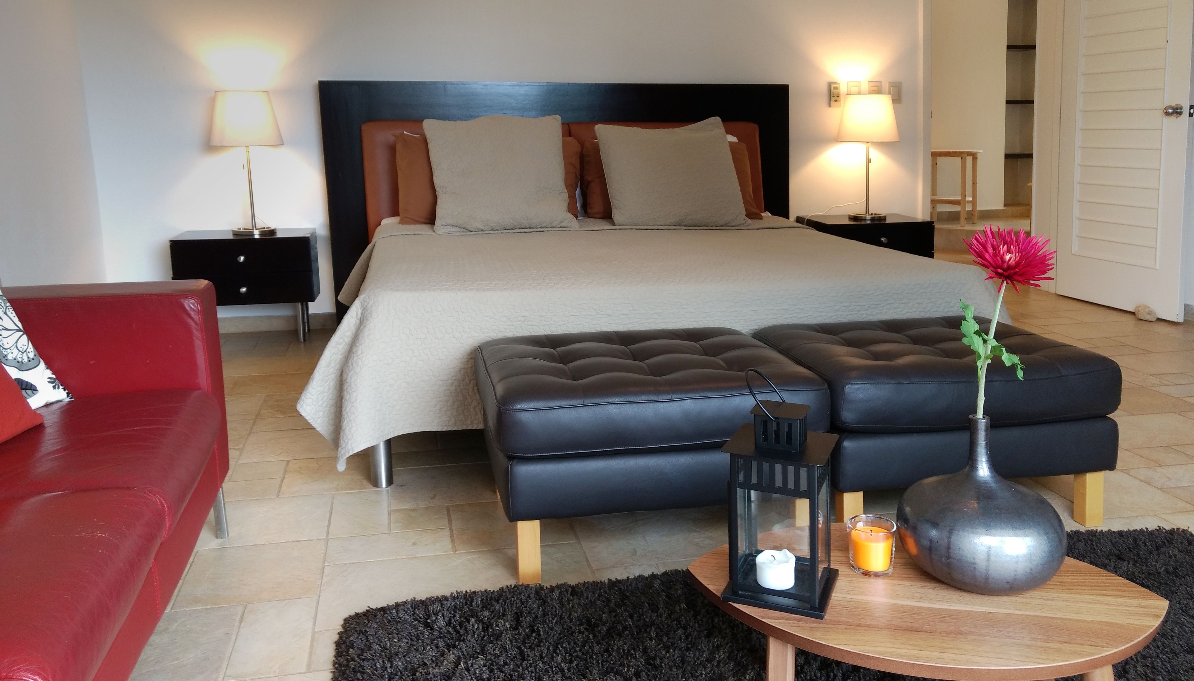 A neatly arranged bedroom with a bed, a red sofa, a black ottoman, a small table with a vase and candle, and two bedside lamps.